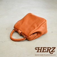 橘色皮革手提包，方形口金設計，帶有單個提把，包身柔軟，置於灰色紋理地面上。