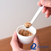 一只手拿著一把不鏽鋼小勺，勺中盛有少量碎塊狀的食物，正準備放入一個白色圓形小碗中。小碗內裝有同款碎塊狀食物。