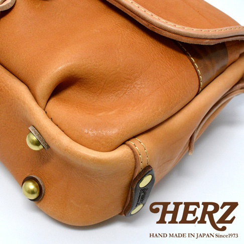 職人手作皮革鹿角扣繩郵差包，呈現原色皮革的自然紋理與光澤，包身設有金屬圓釘裝飾，搭配深棕色皮革標籤，展現簡約復古風格。