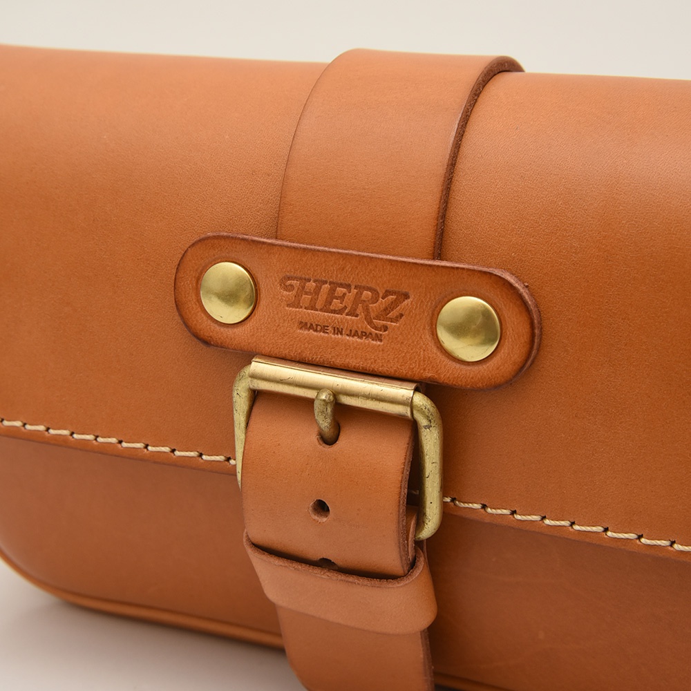 職人手作皮革單扣經典斜背包，以鞣製的淺棕色皮革製成，配有黃銅色金屬扣環和鉚釘，中央皮革片壓印有「HERZ」及「MADE IN JAPAN」字樣，縫線清晰可見。