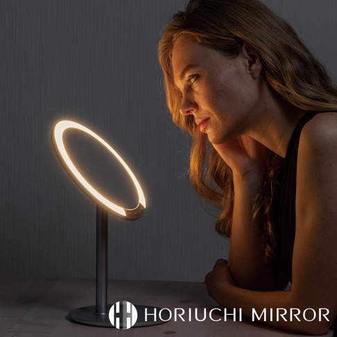 銀色金屬底座的圓形LED化妝鏡，鏡面環繞著暖黃色燈光，一位女性正在鏡前使用。
