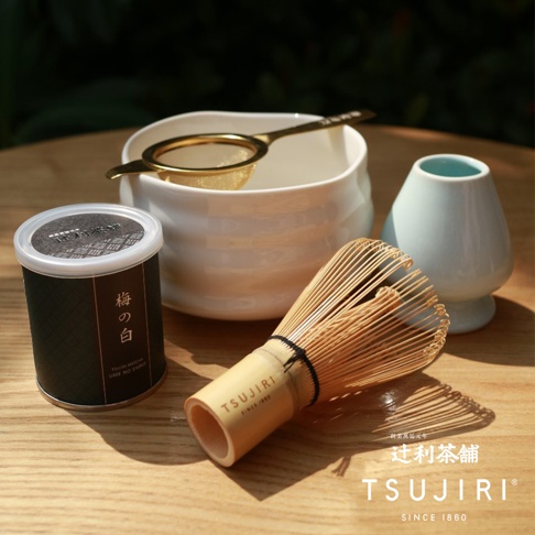 一套抹茶茶具，包含竹製茶刷、白色陶瓷茶碗、金色濾網及淺藍色茶筅架，陳列於木製桌面上。