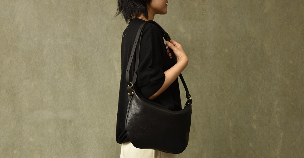 黑色皮革製成的半月形單肩包，由模特兒斜肩背著，包包呈現自然垂墜的質感。