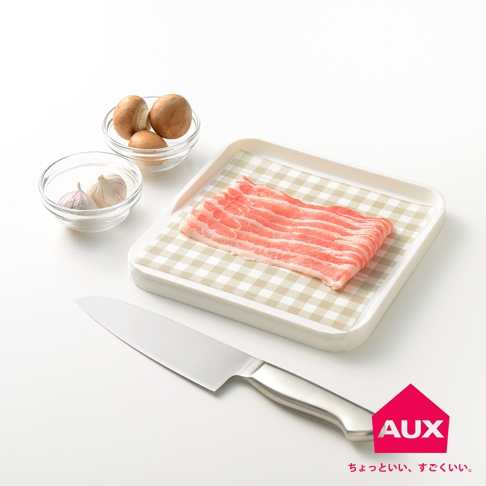 白色迷你砧板上鋪有米白格子紋理的防汙紙，上面擺放著幾片粉紅色生豬肉片。旁邊有裝有蘑菇和蒜頭的透明玻璃碗，以及一把銀色金屬廚刀。