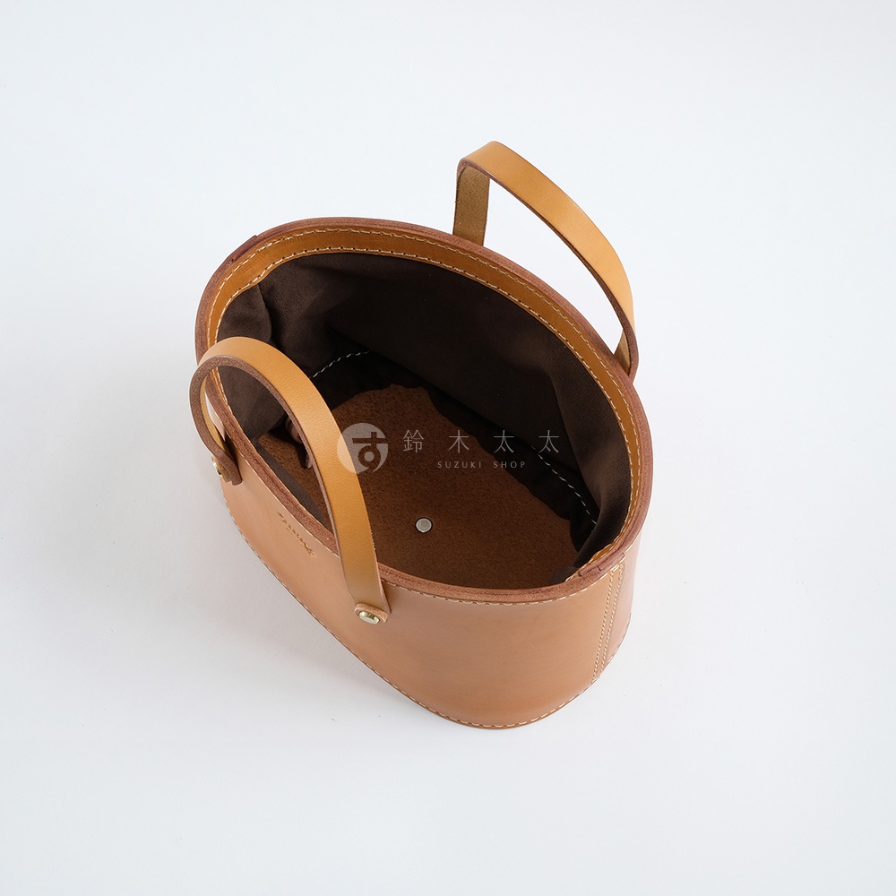 淺棕色皮革束口手提包，以職人手作工藝製成，擁有簡潔的橢圓形輪廓與兩條皮革提把。包內襯為深棕色布料。