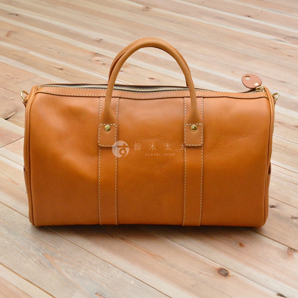焦糖色皮革製成的圓筒形波士頓包，配有雙提把、拉鍊開合和側面掛環，縫線明顯。