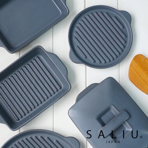 多個深灰色耐熱陶製烤盤，包含圓形及長方形，表面有烤紋，其中一個烤盤帶有蓋子，旁邊有一塊木製鍋鏟。