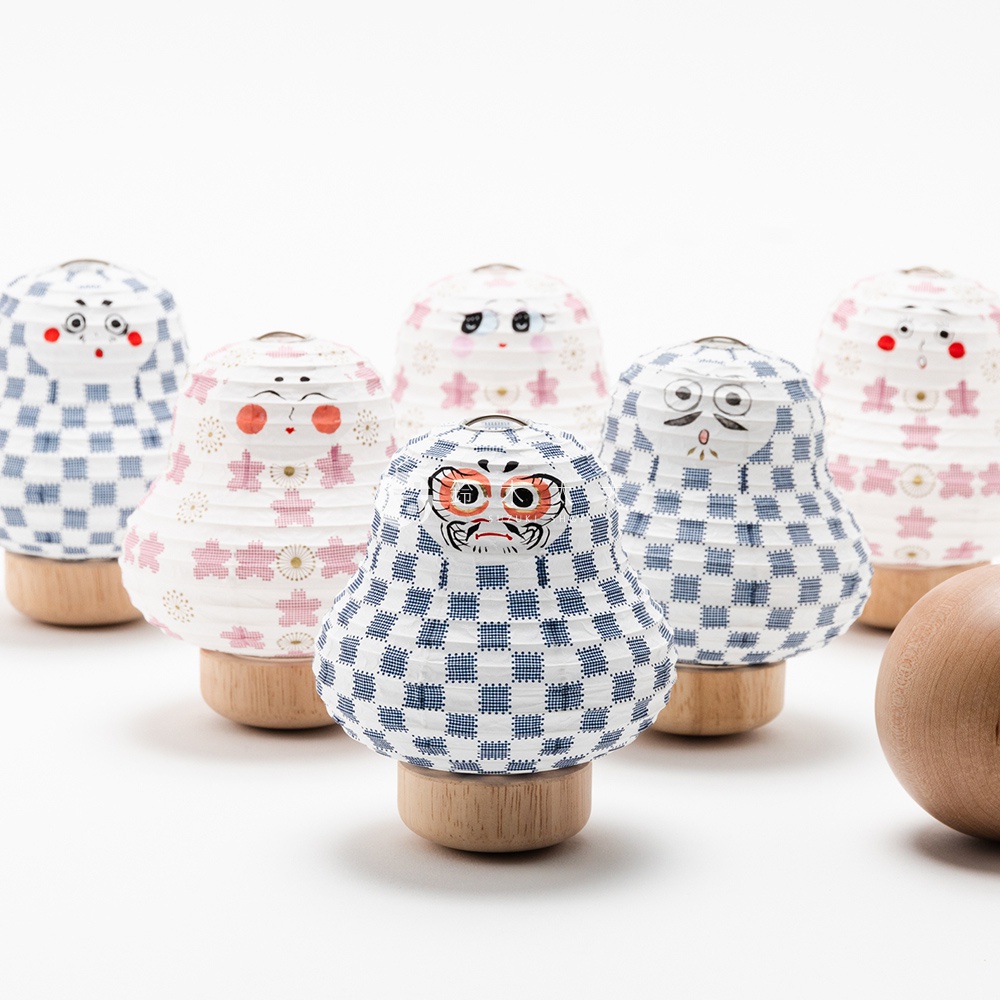 六個達摩不倒翁造型的迷你紙燈籠，以木製底座支撐，燈籠表面印有藍色格紋或粉色櫻花圖案，並繪有不同的臉部表情，呈現日式傳統裝飾風格。