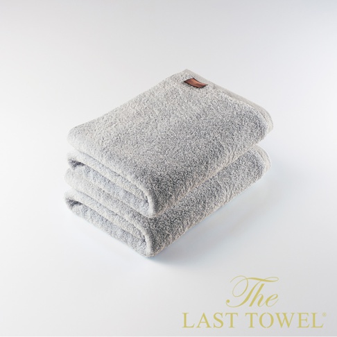 兩條疊放的淺灰色純棉浴巾，帶有棕色皮革標籤，背景為白色。