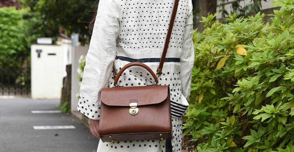 日本HERZ職人手作棕色皮革多功能單肩包，搭配圓形金屬旋轉鎖扣與圓弧形提把，包款旁可見綠葉植物