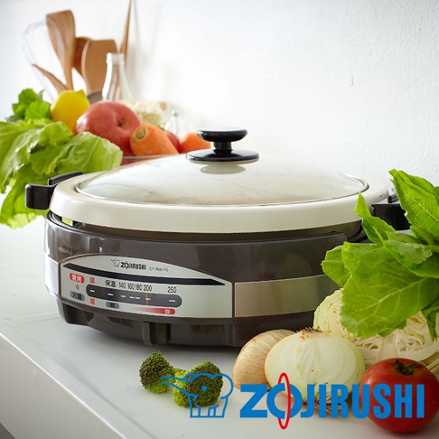 象印土鍋風鐵板萬用鍋 EP-RAF45，鍋身為深棕色與米白色，搭配透明玻璃鍋蓋，鍋蓋中央有黑色把手。鍋身側邊有顯示溫度與功能的控制面板，周圍擺放著新鮮的蔬菜水果，如高麗菜、花椰菜、番茄、蘋果、檸檬和胡蘿蔔。