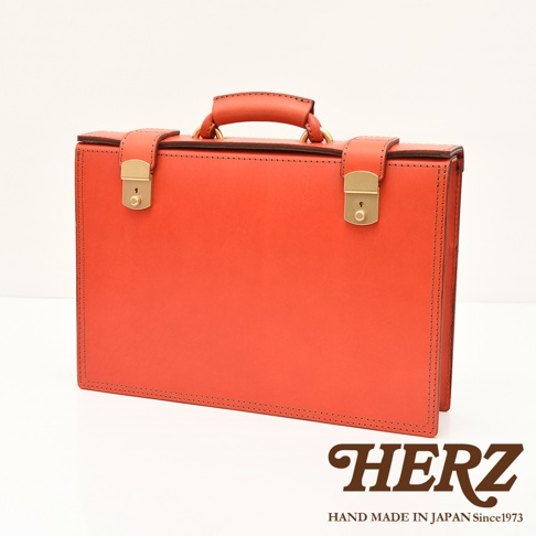 職人手作皮革機師箱 BC-31，橘紅色皮革，設有兩個金色鎖扣和一個提把，箱體邊緣有縫線裝飾。