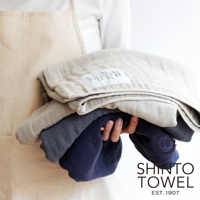 手持三條不同顏色的2.5層紗浴巾，包含淺米色、深灰色和海軍藍。浴巾材質柔軟，可見細緻的織紋。