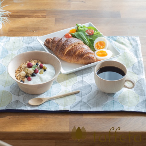 淺色木紋咖啡杯，盛有深棕色咖啡，杯旁擺放著木碗裝的優格麥片、羊角麵包、水煮蛋和沙拉。