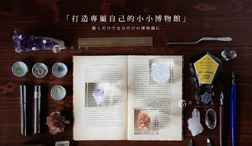 木質桌面陳列著一本打開的舊書，書頁上有德文文字。書本上擺放著數個透明壓克力方塊，每個方塊內嵌有不同顏色的礦石或水晶，包括紫色、白色和橘紅色。周圍散落著其他物品，如金屬鑷子、黃銅尺、鋼筆、墨水瓶、鑽石形狀的裝飾品，以及幾個裝有小礦石的圓形金屬盒。