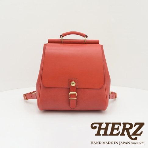 一紅色皮革後背包，包身線條硬挺，帶有屋瓦造型翻蓋，以金屬釦環及皮帶固定，頂部有提把，兩側有可調節的皮革背帶。