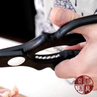 一隻手正握著一對黑色手柄的不鏽鋼萬用料理剪刀，剪刀的刀刃部分有鋸齒狀，靠近手柄處有一個圓形白色標誌。