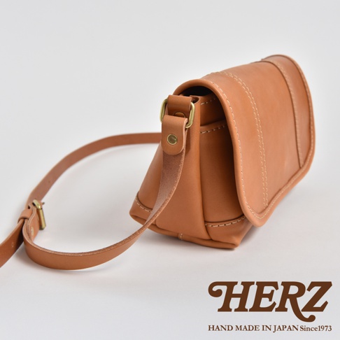 職人手作皮革復古郵差包，採用天然棕色皮革製成，包身線條簡潔，帶有精緻縫線與金屬釦環，可調整長度的皮革背帶。