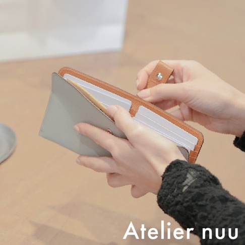 一名女性的手正在開啟 Atelier nuu 的 noble 皮革雙折長夾，錢包內側為淺灰色，外側及扣環處選用棕色皮革，細緻的皮革紋理清晰可見，並展現出其多層卡片收納的設計細節。