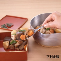 不鏽鋼分食匙盛著多種燉煮食材，包含蓮藕、紅蘿蔔、香菇、竹筍、芋頭等，食材呈現天然色澤，部分紅蘿蔔切成花形。分食匙傾斜，食材部分落入一旁紅漆木盒中。