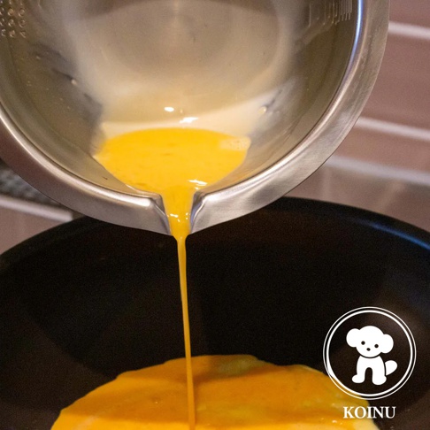 不鏽鋼料理盆傾斜，將金黃色液體（可能是蛋液）倒入黑色鍋中。盆內有刻度標記。