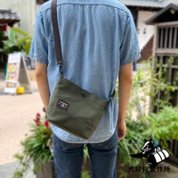 一個身穿牛仔襯衫的男人，將一個墨綠色純棉帆布迷你側背包斜挎在肩上。包包上有一個方形的紅色與黑色標籤，標籤上有日文文字和一個黑白狗的圖案。