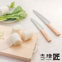 兩把銀色刀刃、淺木色手柄的萬用刀（Santoku knife），一把放在白色木紋背景上，另一把放在淺木色砧板上，砧板上有切好的白色塊狀蔬菜，以及一盤綠葉蔬菜和白蘿蔔。