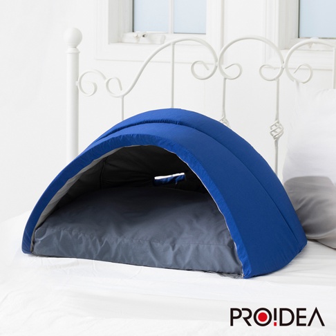 藍色圓拱形遮光舒眠罩，內有灰色軟墊，頂部有通風孔，罩身為布料材質，放置於白色床鋪上。