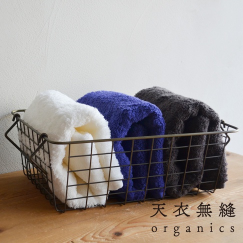 三條捲起的毛巾，分別為白色、藍色和炭灰色，整齊擺放在金屬絲籃中，毛巾材質蓬鬆柔軟。