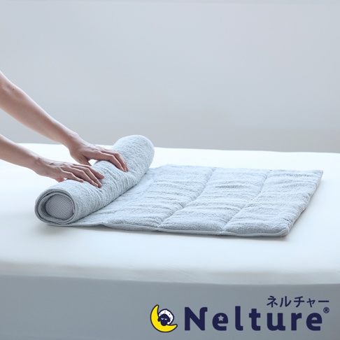 一隻手正在將一條淺灰色的毛巾枕捲起，毛巾枕鋪在白色的床墊上，枕頭上有縫線分隔的格子，右下角有 Nelture 品牌標誌，標誌為一彎月亮上有隻綿羊，旁邊是日文「ネルチャー」及英文「Nelture」。