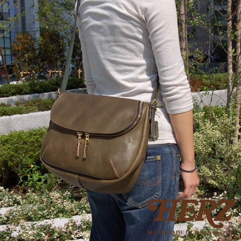 一位女士身穿淺色長袖上衣和牛仔褲，背著一個綠褐色皮革斜背包，包包上有兩個拉鍊，背景是綠意盎然的戶外景觀，右下角有HERZ品牌標誌。