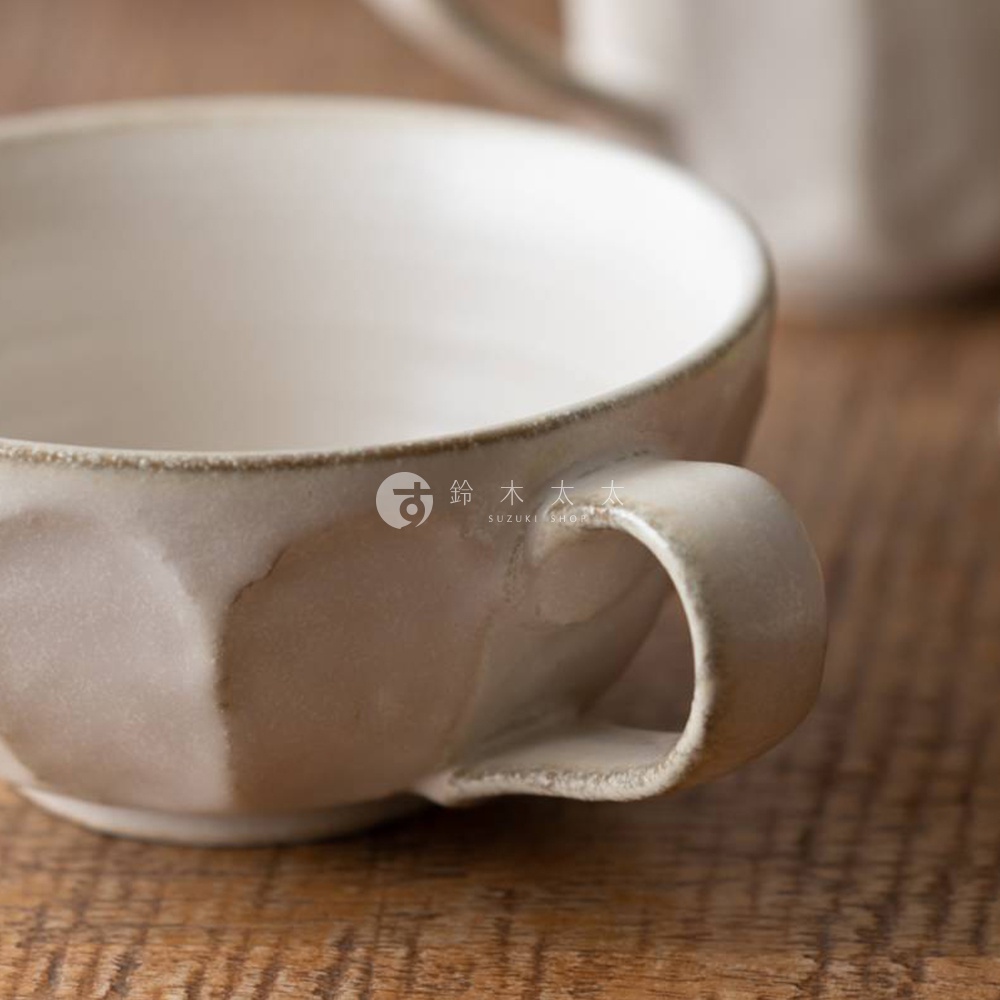 米白色的美濃燒湯杯碗，帶有簡約的把手，碗身呈現多邊形凹凸紋理，表面有自然的釉色變化，散發樸實的質感。