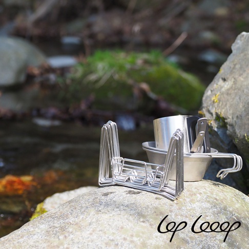 銀色不鏽鋼伸縮式瀝水架，結構層疊，附帶一個銀色不鏽鋼杯和夾子，置於岩石上，背景是溪流與綠色植被。