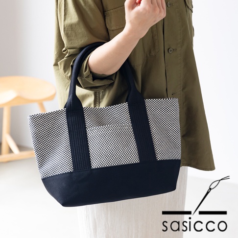 一款黑色與白色格紋刺子織迷你托特包，配有黑色織帶提把與底部，由一位身穿卡其色上衣的人士手提。