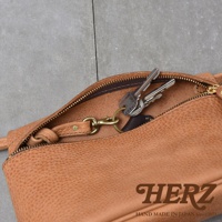 HERZ 品牌標誌，下方印有 HAND MADE IN JAPAN Since1973 字樣，一條棕色皮革斜背包的拉鍊打開，露出內側，並掛有鑰匙串。