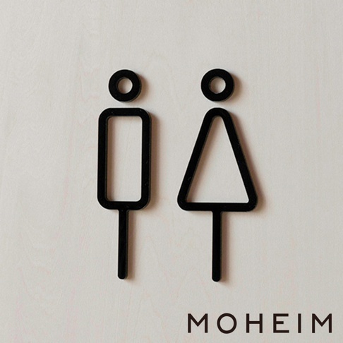 一對黑色簡約男女廁所標示牌，分別為男性（矩形身體）和女性（三角形身體）圖案，皆有圓形頭部和長形腿部，立體設計，材質疑似為壓克力或木材，背景為淺色紋理表面。