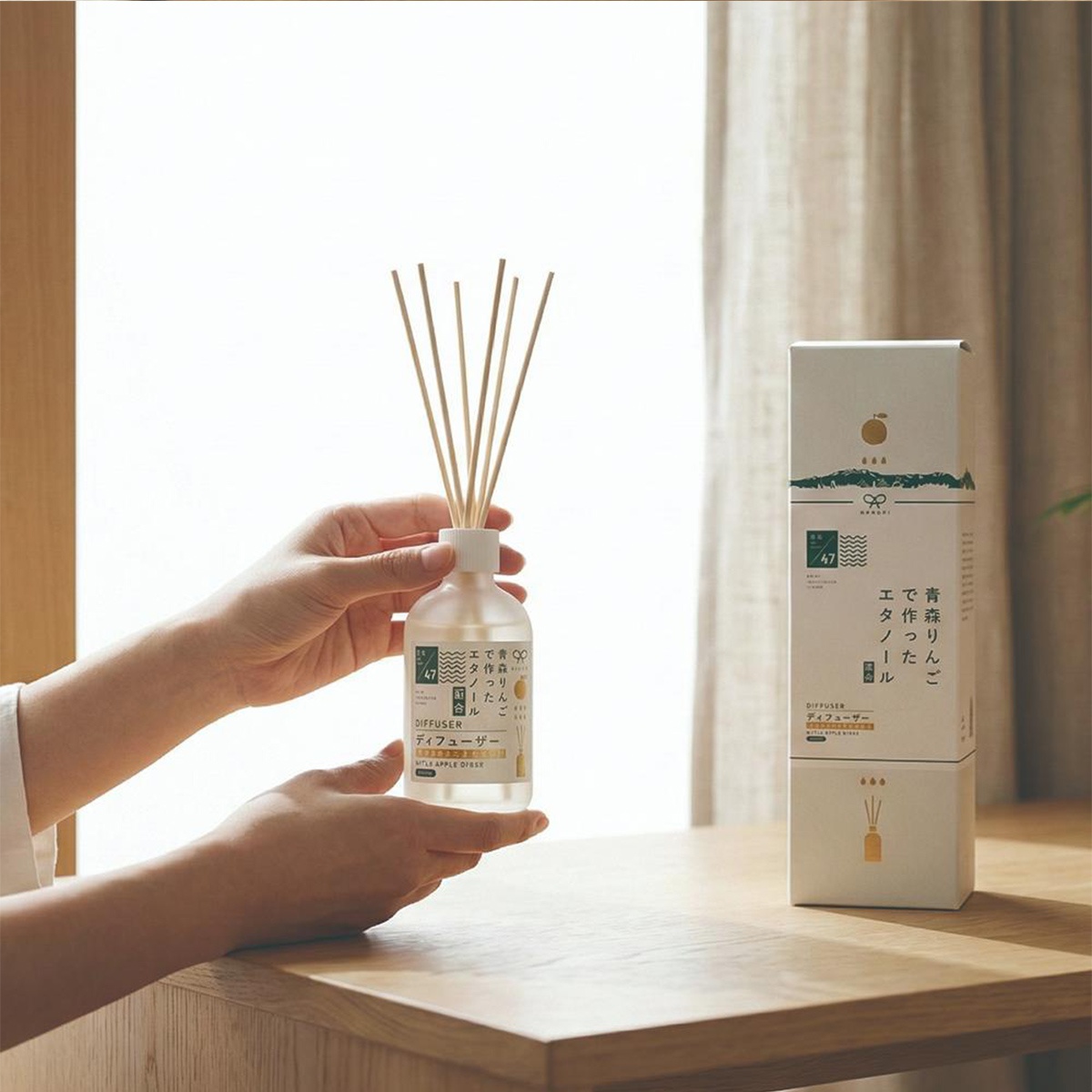 兩隻手捧著一瓶透明擴香瓶，瓶內裝有淡黃色液體，瓶口插著幾根擴香竹，旁邊擺放著同款商品紙盒。