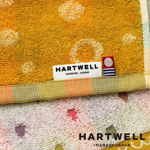 這是一張展示 Hartwell 今治毛巾細節的特寫照片，毛巾為鮮豔的黃色，表面帶有圓形點狀織紋，底部邊緣裝飾著紅白相間的格紋滾邊。標籤上清楚印有 HARTWELL IMABARI JAPAN 的字樣，旁邊還附有一個紅白藍色的圓形圖騰小標籤，整體質感細膩精緻。