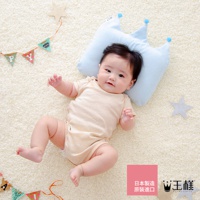 淺藍色嬰兒皇冠枕，枕頭上有兩顆藍色絨球裝飾，材質柔軟，適合0-6個月嬰兒使用，搭配條紋連身衣的嬰兒躺在白色絨毛地毯上，背景有彩旗和星星裝飾。