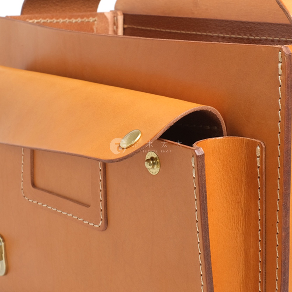 職人手作皮革橫式書包的局部特寫，展示了淺棕色皮革材質、金屬扣環以及手工縫線細節。