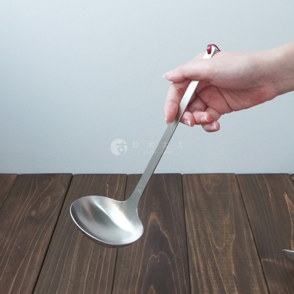 一只手正握著不鏽鋼萬用湯勺，湯勺為銀色，勺身呈現圓弧形，手柄細長，末端有紅色裝飾。