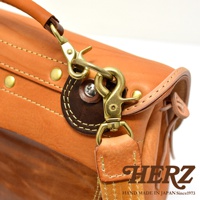 鞣製皮革製成的郵差包，帶有鹿角狀扣環和鉤扣，搭配皮革提把和肩帶，整體為淺棕色，細節處使用深棕色皮革點綴，並以縫線和金屬配件強化結構。