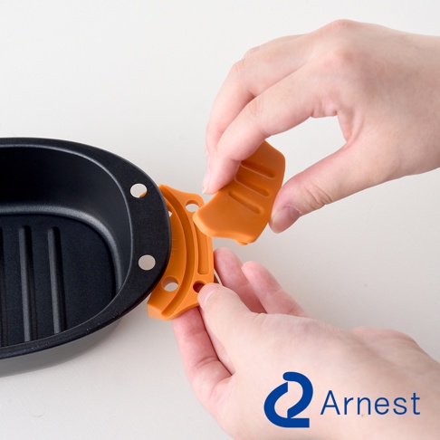Arnest 品牌 logo 下方，雙手正將橘色配件組裝到黑色煎鍋邊緣，鍋具內部有條紋。