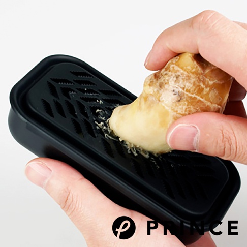 一隻手正將薑塊在黑色兩用盒的磨泥槽上磨擦，盒身為塑膠材質，槽內有菱形紋路。