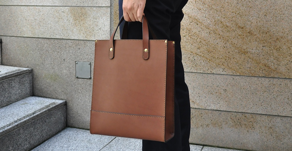 棕色皮革直式托特包，配有雙提把和金色鉚釘裝飾，皮革表面光滑，有縫線細節。