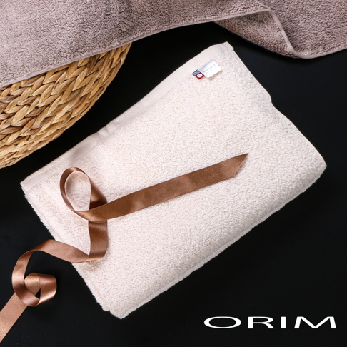 一條米白色毛巾，材質柔軟，上面繫著一條銅色緞帶。毛巾一角有品牌標籤。背景為黑色，左上方有一籃子裝著的灰色毛巾。