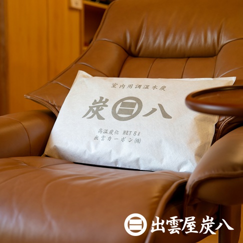 白色包裝袋，上面印有灰色日文「炭」字及「八」字標誌，寫有「室內調濕木炭」、「高溫炭化 NET 8L」等字樣，置於棕色皮質沙發上。