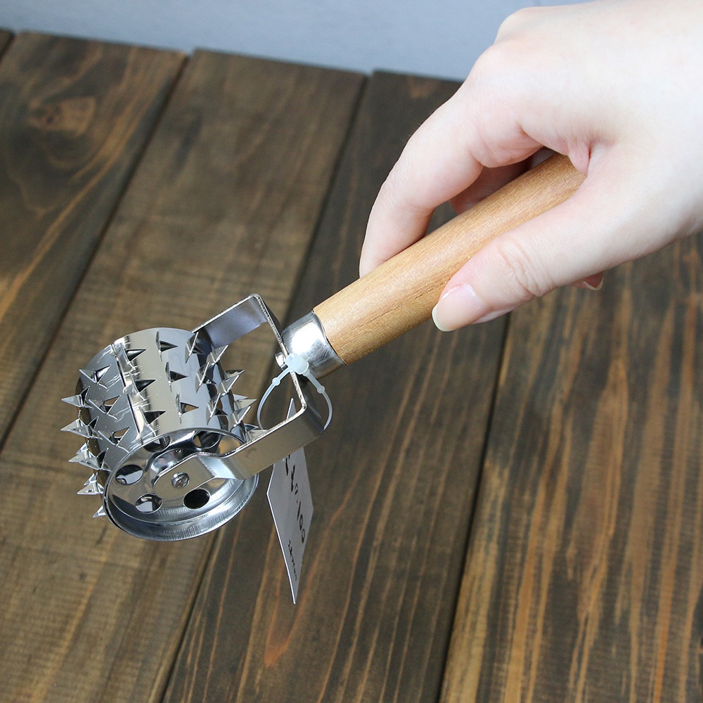 一只手握著不鏽鋼滾輪斷筋器，其滾輪部分設有多個尖刺，連接木製手柄，背景為深色木紋桌面。