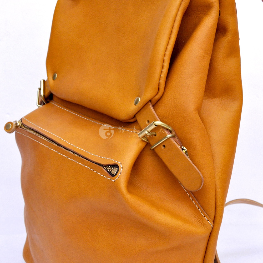 職人手作皮革後背包，顏色為暖棕色，具有多口袋設計，包含一個拉鍊前袋和翻蓋式主袋，袋蓋上飾有金屬釦和皮帶扣，整體材質為柔軟的皮革，車線為白色。
