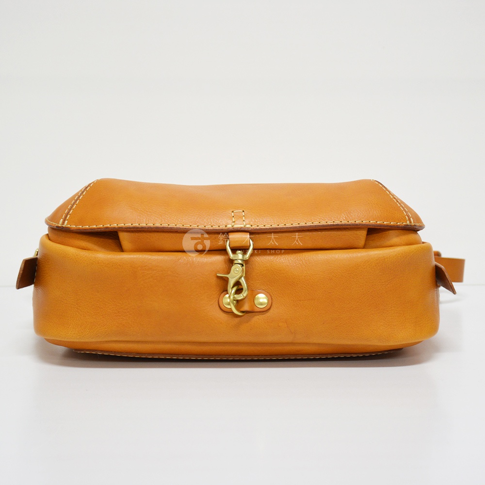 職人手作皮革復古翻蓋郵差包，以溫潤的淺棕色皮革製成，設計簡潔，配有金屬鉤扣與鉚釘，展現經典復古風格。